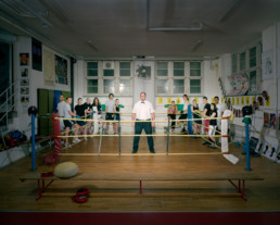 Inszeniertes Gruppenbild von Mitglieder von einem Boxclub in einem Boxkeller. Der Ringrichter steht alleine im Ring, die Boxer stehen um den Ring.