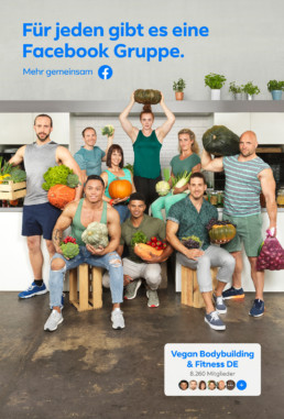 In einer hellen, modernen Küche stehen Bodybuilder und Bodybuilderinnen umgeben von Früchten und Gemüsen in ihrer Bodybuildingbekleidung zusammen und posieren zur Kamera.