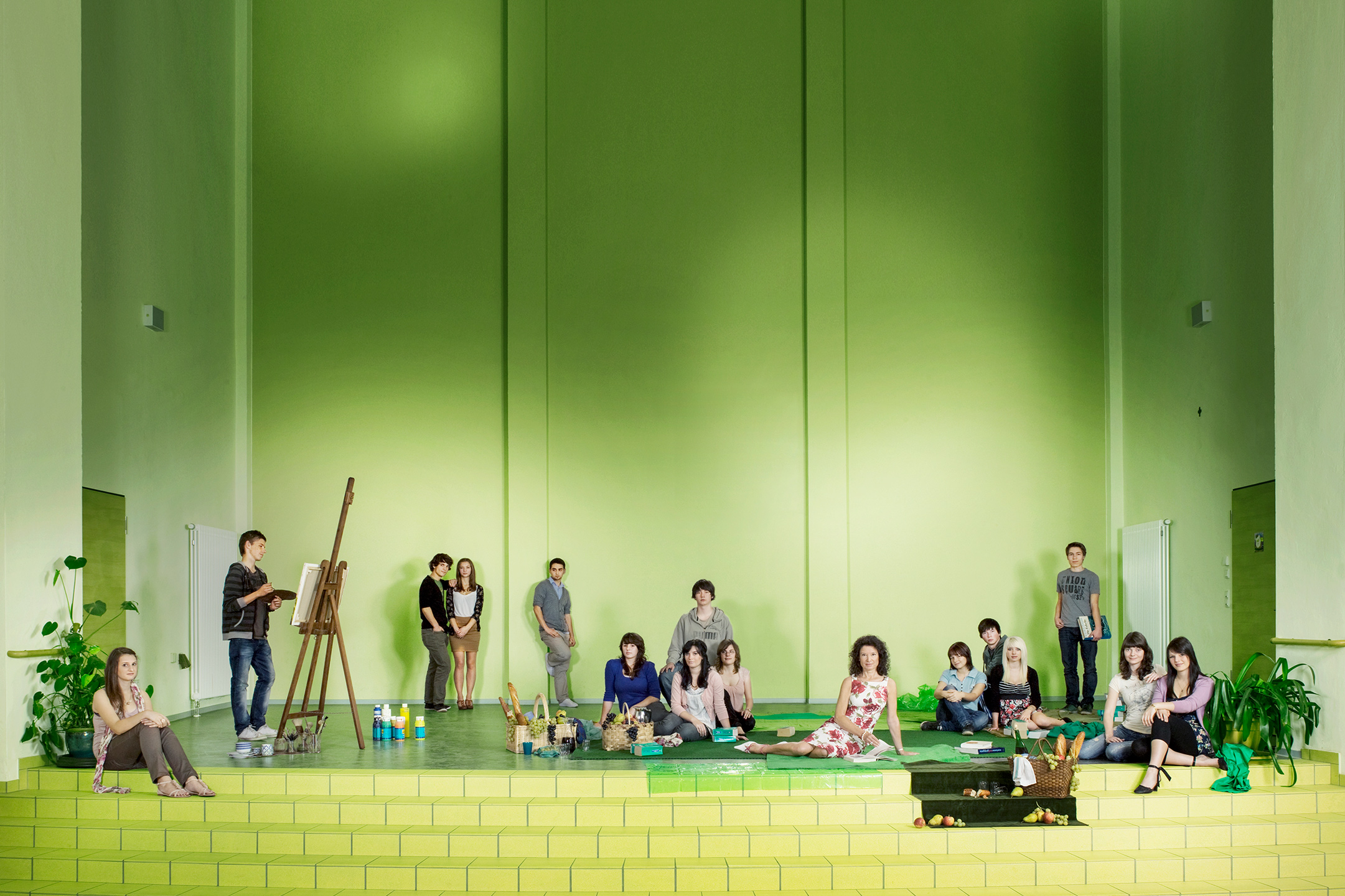 uf der Bühne einer Schulaula sitzen und stehen alle Schüler und Schülerinnen wie in einem modernen Bild Déjeuner nur l’Herbe herum. Mit Essen, Zeichen und Staffelei, Bücher, … Die Lehrerin sitzt entspannt auf einer Pic-Nic-Decke