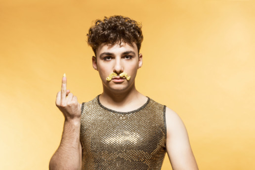 Ein junger Mann mit einem goldenen Muskle-Shirt zeigt uns den Stinkefinger. Er hat sich mit Senf einen Schnauz gemalt. Der Hintergrund ist gelb.