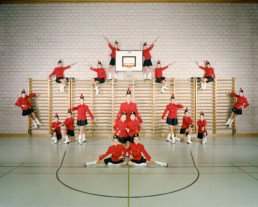 Inszeniertes Gruppenbild von Mitglieder einer Showtanzgruppe, welche in ihrer Mjorettenuniform-Kleidung einer Turnhalle symmetrisch vor einer Sprossenwand angeordnet sind.