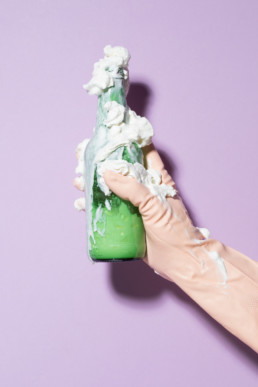 Eine Hand in einem hautfarbenen Plastikhandschuh hält vor einem lila Hintergrund eine Bierflasche, aus welcher viel Rasierschaum quillt