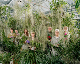 Inszeniertes Gruppenbild von Mitglieder*innen eines Orchideenclub. Alle stehen, dicht umgeben von vielen Orchideen und Pflanzen, in einem Gewächshaus.