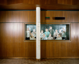 Inszeniertes Gruppenbild von Mitglieder eines Pfeifen-rauch- Vereins. Hinter Glas in einem abgetrennten kleinen Raum sitzen und stehen die Männer in ihrem Vereinshemden und rauchen Pfeife.