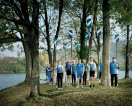 Inszeniertes Gruppenbild von Mitglieder*innen eines Ruderclubs. Sie stehen in ihrer Vereins-Ruder-Kleidung am Ufer von einem Fluss zwischen den Bäumen. Jede/r hat ein Riemen in der Hand.