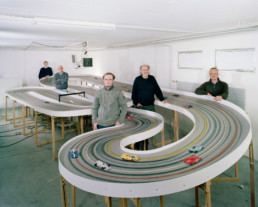 Inszeniertes Gruppenbild von Männer eines Slot-Car-Racing-Clubs. In einem Keller habe sie ihre Modellautorennbahn aufgebaut und stehen ringsum.