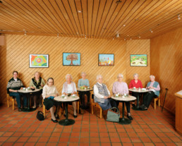 nszeniertes Gruppenbild von meist älteren Damen, welche in einem getäferten Raum verteilt mit Ihren Strickarbeiten an kleinen Tischen sitzen.