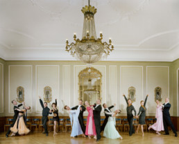 Inszeniertes Gruppenbild von Standardtänzerinnen und -Tänzer in ihren schicken Tanzkleider in einem edlen Ballsaal mit einem grossen Leuchter.