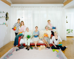 Inszeniertes Gruppenbild von Mitgliederinnen eines Tupperware-Clubs. Die Damen sitzen alle auf einem Sofa in einem hellen Wohnzimmer. Auf Ihren Knien haben sie Tupperware-Produkte.