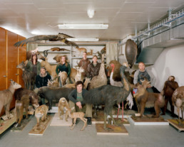 Inszeniertes Gruppenbild von Mitglieder eines Präparatorenverbands. In einem Keller-Archiv sitzen und stehen die Präparatorinnen mitten zwischen vielen präparierten Tieren.