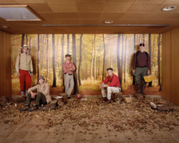 Inszeniertes Gruppenbild von Mitglieder von einem Pilzkunde-Verein. Die Männer und Frauen stehen in einem braunen Raum vor einer Herbstwaldtapete. Am Boden viel Laub und Pilze.