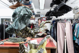 In einem Raum von einer Kleidersammelstelle hängen viele Kleider. Eine Mitarbeitern steht zwischen den Hosen, Hemden und Blusen.