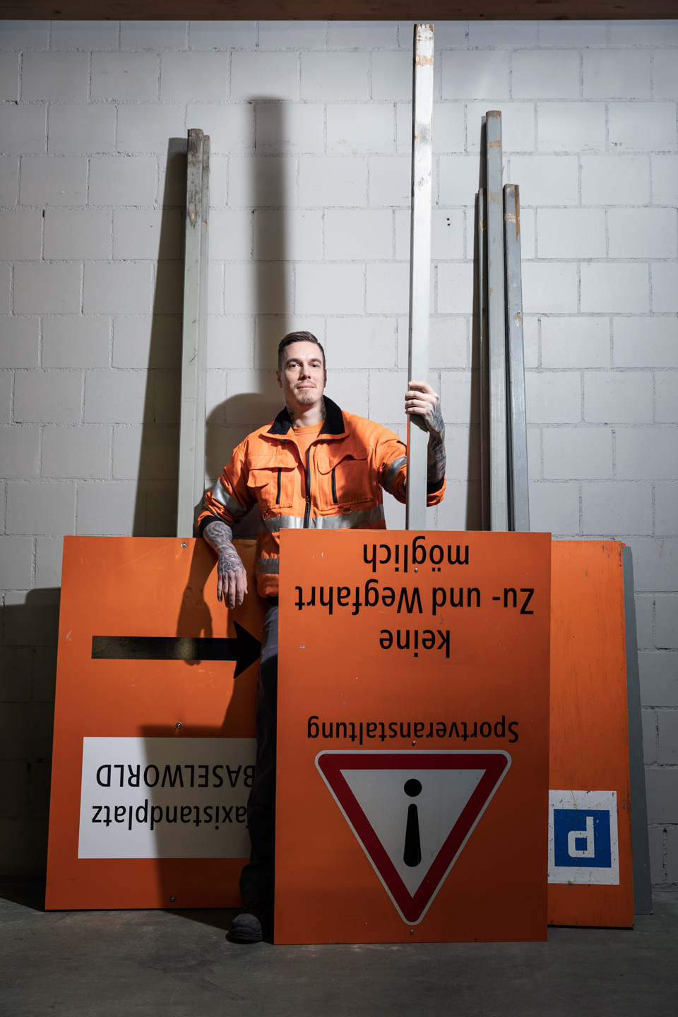 An einer grauen Backsteinwand sind einige grosse Strassenschilder angelehnt. Dazwischen steht ein Mitarbeiter.