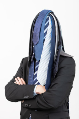 Ein Mann steht vor einem hellen Hintergrund. Unzähligen Krawatten liegen auf seinem Kopf und verdecken sein Gesicht.