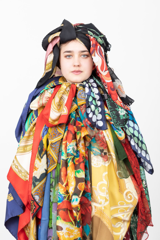 Vor einem hellen Hintergrund steht eine Frau. Sie ist mit verschiedensten Foulards überhäuft.